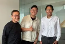 Three smiling men pose indoors against a neutral background.