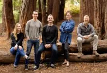 Five people pose outdoors, sitting and standing on a fallen tree trunk.