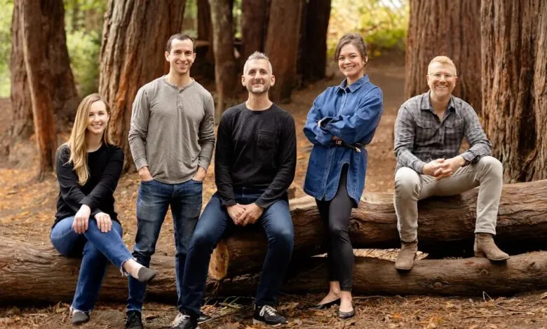 Five people pose outdoors, sitting and standing on a fallen tree trunk.