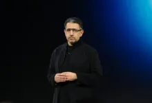 Man with glasses in black coat stands against a blue backdrop.