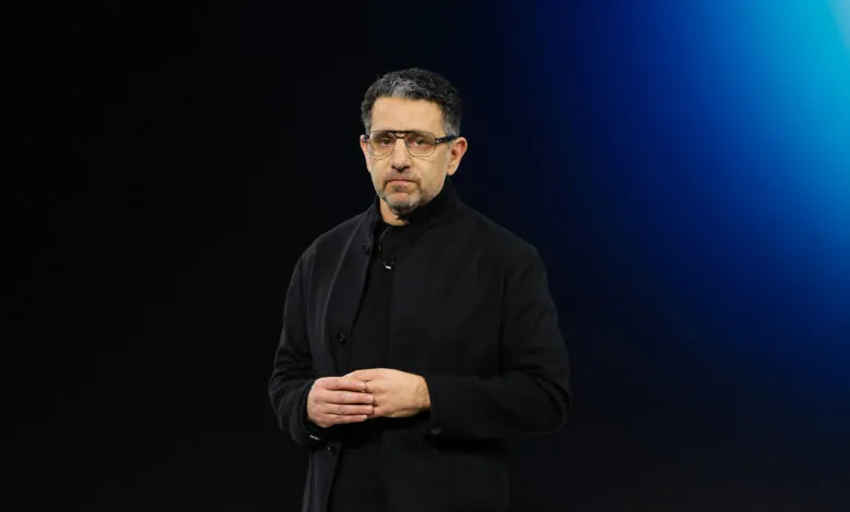 Man with glasses in black coat stands against a blue backdrop.