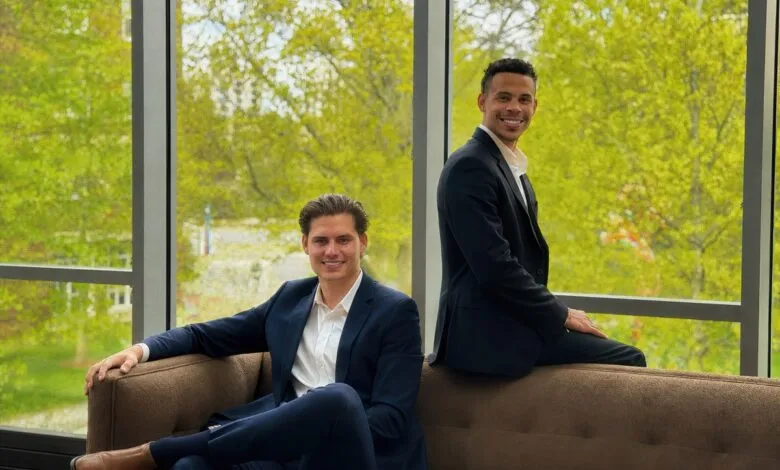 Two men in suits pose on a couch in front of a large window.