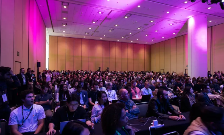 Large crowd of people seated at a conference, bathed in pink light.