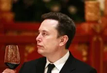Elon Musk in suit holding glass of red wine, looking thoughtful.
