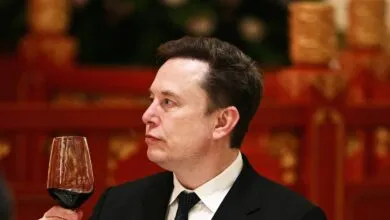 Elon Musk in suit holding glass of red wine, looking thoughtful.