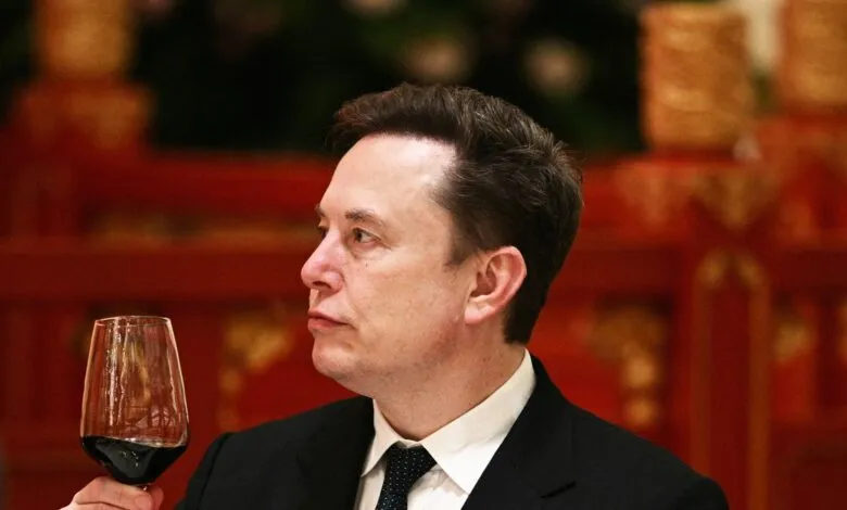 Elon Musk in suit holding glass of red wine, looking thoughtful.