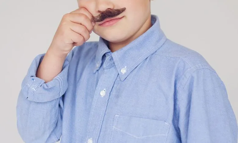 Young boy wearing a fake mustache and a blue button-down shirt.
