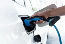 Hand plugs blue charging cable into white electric car port.