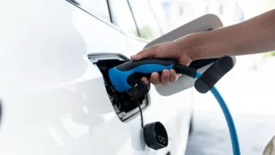 Hand plugs blue charging cable into white electric car port.