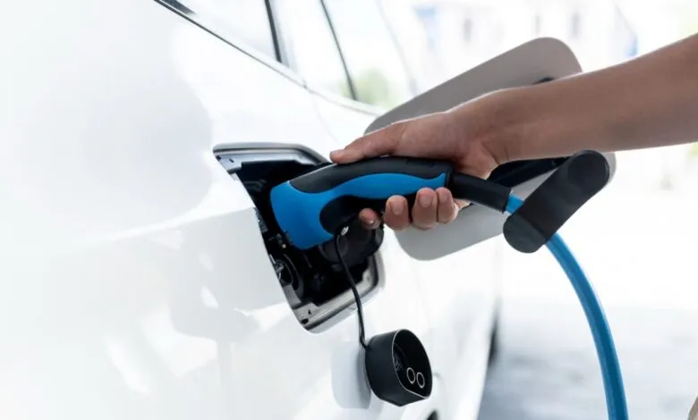 Hand plugs blue charging cable into white electric car port.