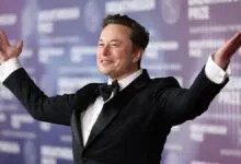 Elon Musk in tuxedo with arms raised on red carpet.