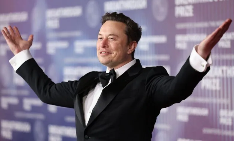 Elon Musk in tuxedo with arms raised on red carpet.