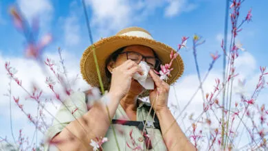 Woman in hat uses tissue, likely experiencing allergy symptoms outdoors.
