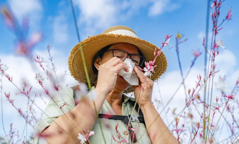 Woman in hat uses tissue, likely experiencing allergy symptoms outdoors.