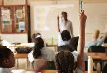 Students in classroom with raised hands, teacher at whiteboard.