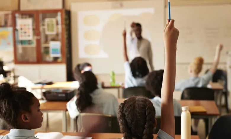 Students in classroom with raised hands, teacher at whiteboard.