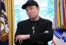 Elon Musk with arms crossed, wearing a black baseball cap.