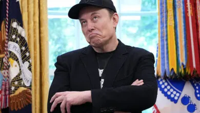Elon Musk with arms crossed, wearing a black baseball cap.