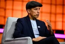Man in suit and cap sits in chair, speaking with microphone.