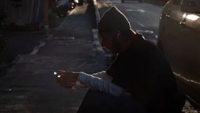 Man in beanie sits on curb using phone, bathed in sunlight.
