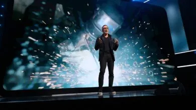 Man on stage presenting with welding sparks on large screen behind him.