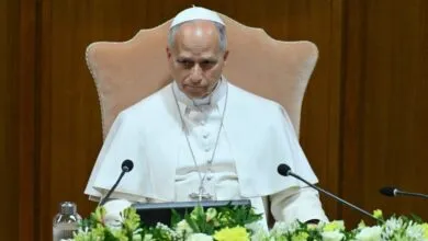 Pope Leo XIV seated at a table with microphones and floral arrangements.