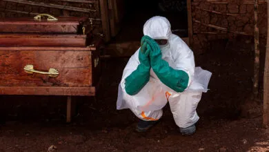 Health worker in PPE crouches near coffin, hands clasped in prayer.