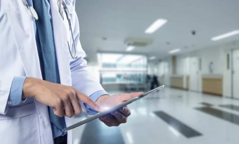 Doctor in lab coat using tablet in hospital hallway.