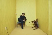 Man sits in yellow room with overturned chair, creating a surreal, unsettling atmosphere.