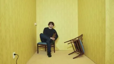 Man sits in yellow room with overturned chair, creating a surreal, unsettling atmosphere.