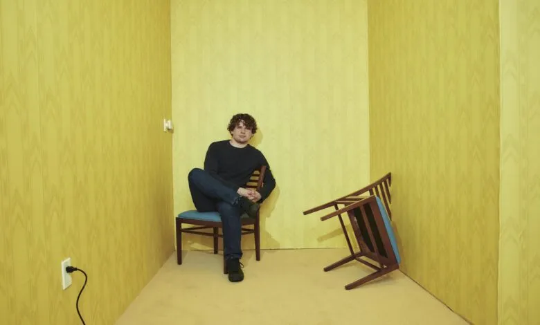 Man sits in yellow room with overturned chair, creating a surreal, unsettling atmosphere.