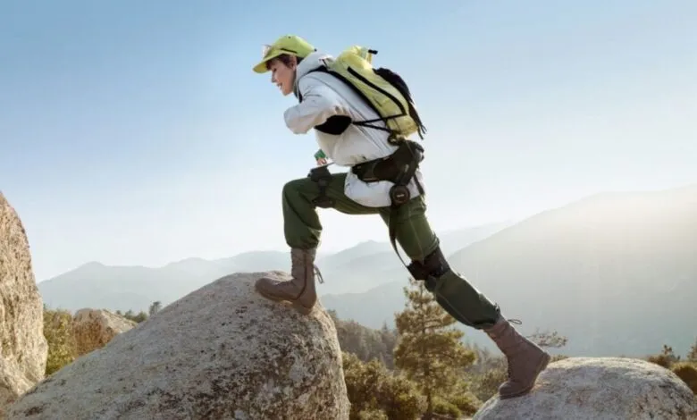 Hiker wearing exoskeleton climbs a rock with mountains in background.