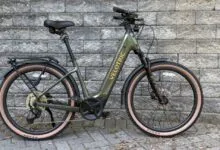 Olive green Velotric e-bike with tan tires against a gray brick wall.