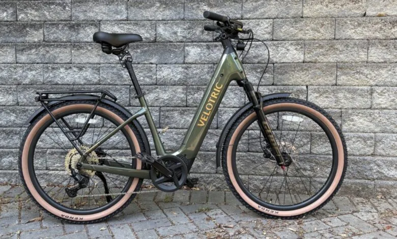 Olive green Velotric e-bike with tan tires against a gray brick wall.