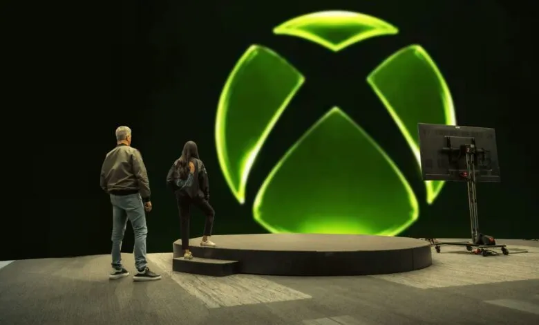Two people stand before a large Xbox logo on a stage.