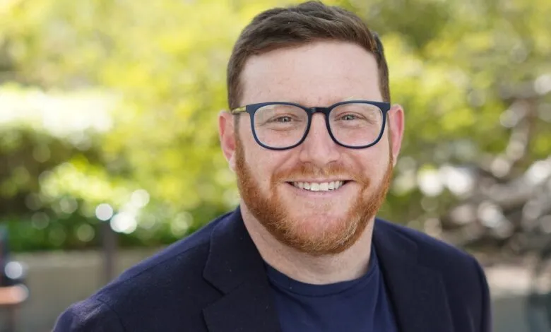 Smiling man with red beard and glasses in front of blurred green background.