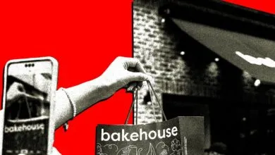 Hand holds Bakehouse bag, phone reflects store, red background.