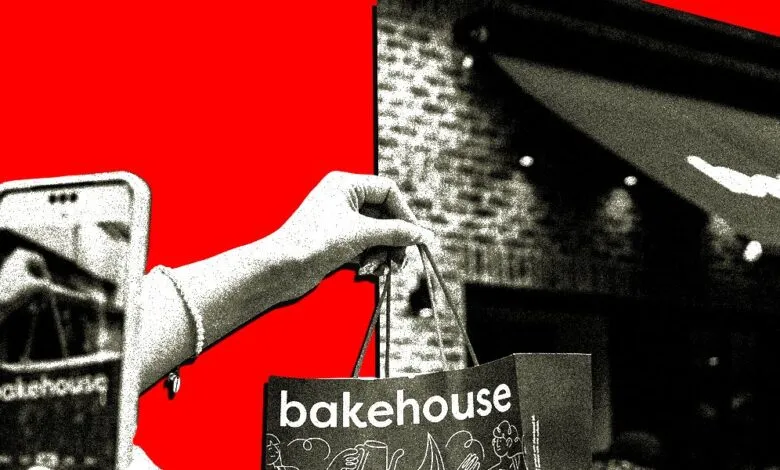 Hand holds Bakehouse bag, phone reflects store, red background.