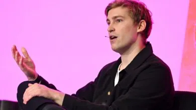 Joe Alwyn speaking on stage with a pink backdrop.
