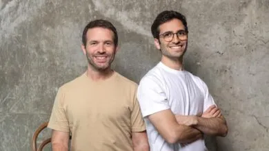 Two smiling men stand side-by-side against a textured gray wall.