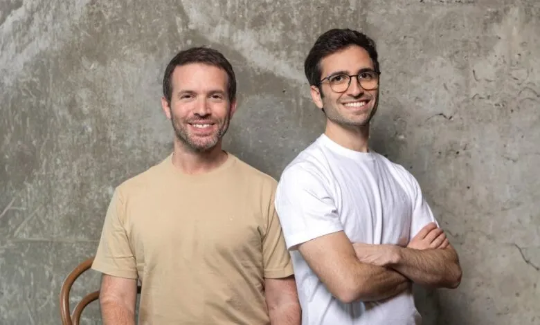Two smiling men stand side-by-side against a textured gray wall.