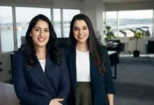 Two smiling women in blazers stand in a modern office setting.