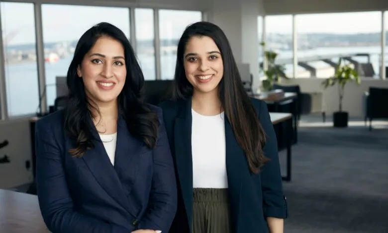 Two smiling women in blazers stand in a modern office setting.