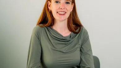 Smiling woman with red hair sits at a white table in front of a gray background.