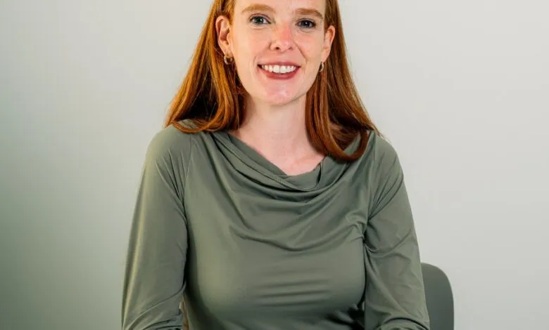 Smiling woman with red hair sits at a white table in front of a gray background.