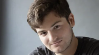 Portrait of a young man with brown hair smiling softly against a gray background.