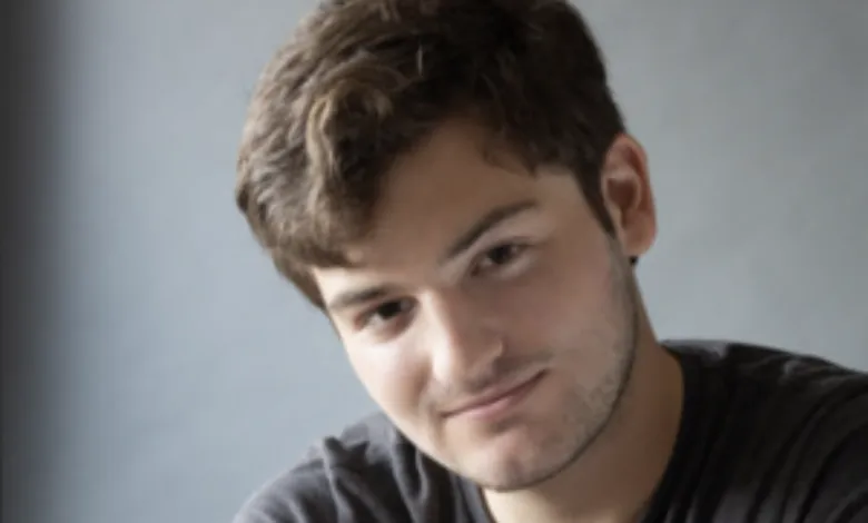 Portrait of a young man with brown hair smiling softly against a gray background.