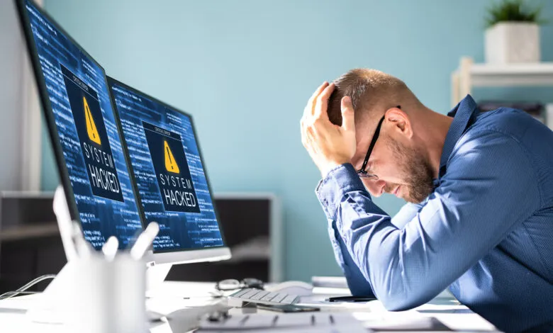 Frustrated man with head in hands, two monitors display 'System Hacked' message.