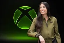 Asha Sharma poses in front of the Xbox logo, smiling and wearing a green jacket.