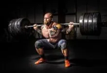 Muscular man with tattoos squats with heavy barbell in gym.